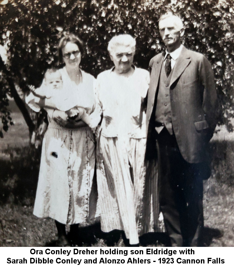 Black and white photo of Ora Conley Dreher with bobbed hair and black rimmed glasses, holding her baby son Eldridge Conley Dreher, standing next to her mother, white-haired Sarah Dibble Conley and grey-haired and -moustached Alonzo Ahlers, on a summer lawn before some trees in Cannon Falls.